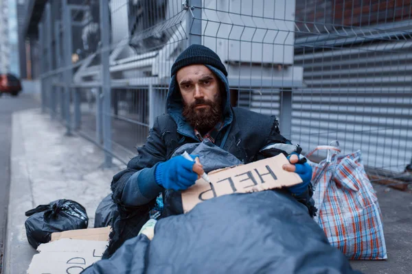 髭を生やした汚れたホームレスは 街の通りにヘルプサインを書きます 貧困は社会問題 ホームレスや孤独 アルコール依存症や飲酒依存症 都市部の孤独 — ストック写真
