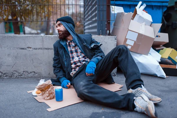 满嘴胡须的肮脏乞丐 带着食物坐在城市街上的垃圾桶边 贫穷是一个社会问题 无家可归 酗酒和酗酒成瘾 — 图库照片