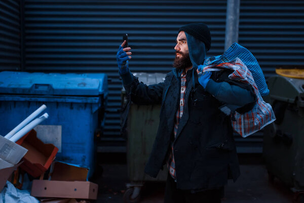 Happy beggar found mobile phone in trashcan on city street. Poverty is a social problem, homelessness and loneliness, alcoholism and drunk addiction, urban lonely