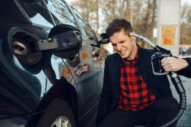 Stressed man with gun at his head on gas station, high price on fuel concept. Petrol fueling, gasoline or diesel refuel service clipart