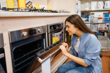 Young woman choosing electric oven in electronics store. Female person buying home electrical appliances in market, housewife looking goods in shop clipart