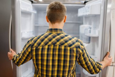 Young man at the opened refrigerator in electronics store. Male person buying home electrical appliances in market clipart