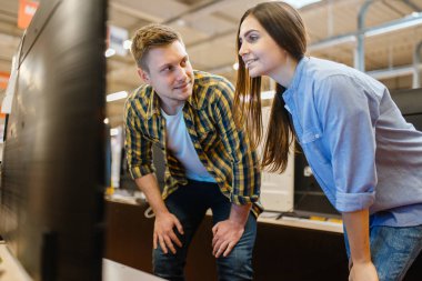 Elektronik mağazasında televizyon izleyen aile çifti. Erkek ve kadın marketten ev aletleri satın alıyorlar.