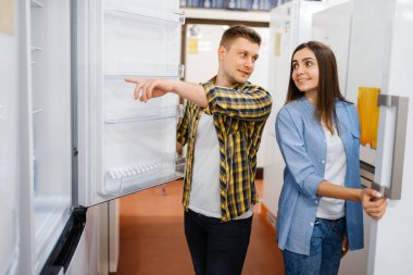 Genç bir çift elektronik mağazasında buzdolabı seçiyor. Erkek ve kadın marketten ev aletleri satın alıyorlar.