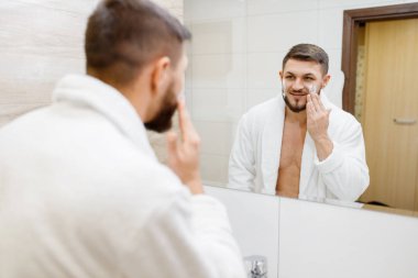 Bornozlu bir adam banyodaki aynada tıraş köpüğü sürüyor. Rutin sabah hijyeni. Lavabodaki erkek kişi deri ve vücut tedavi prosedürlerini gerçekleştiriyor.