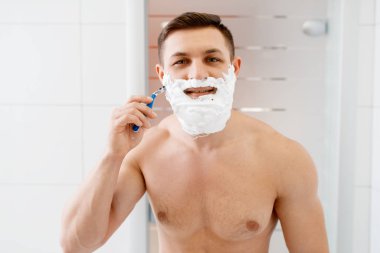 Adam sakalını banyoda tıraş ediyor, sabah hijyeniyle. Erkek kişi deri ve vücut tedavi prosedürlerini gerçekleştirir.
