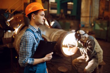 Defterde usta, fabrikada maskeli kaynakçı, fabrikada metal boru ile çalışan bir adam, kaynakçılık yeteneği. Metal endüstrisi, çelik ürünlerinin endüstriyel üretimi