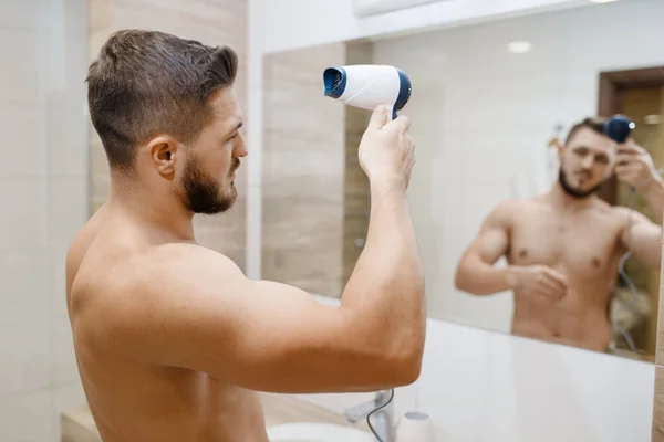 男はヘアドライヤー 朝の衛生手順で彼の髪型を乾燥させます 浴室の鏡の中のアスレチック男性 — ストック写真