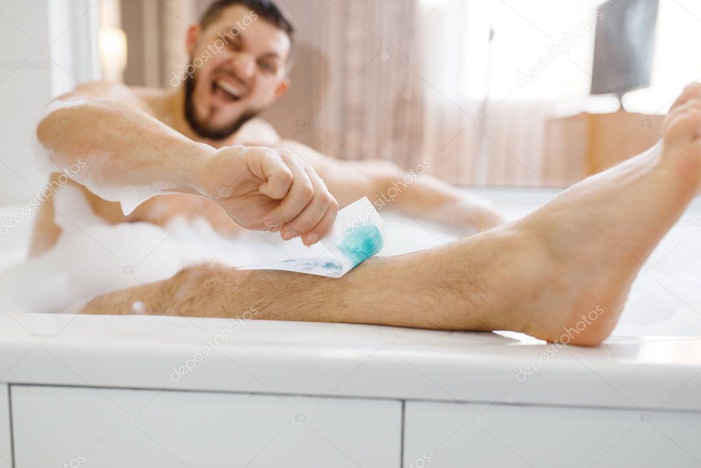 Man removes hair on legs in bath with foam, morning hygiene, waxing depilation. Male person resting in bathroom, skin and body treatment procedures