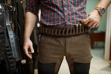 Silah dükkanında avlanmak için mühimmat kemeri ve üniforma seçen bir adam. Silah deposunda ayakta duran avcılar için teçhizat, spor atıcılığı hobisi 