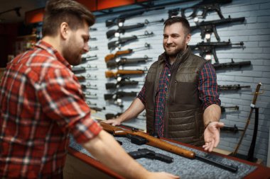 Man and owner choosing rifle in gun shop. Euqipment for hunters on stand in weapon store, hunting and sport shooting hobby  clipart