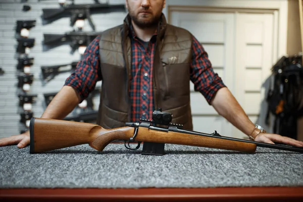 Man with rifle standing at counter in gun shop. Euqipment for hunters on stand in weapon store, hunting and sport shooting hobby