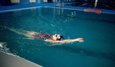 Havuzda sırtüstü yüzen mayo, kep ve gözlüklü bir kadın yüzücü. Kadın suda yüzüyor, spor eğitimi alıyor, hareketli bakış açısı var.