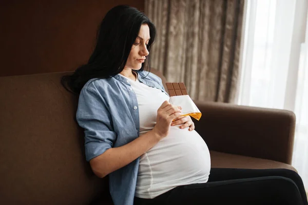 お腹をソファに座りチョコレートを食べる妊婦さん 妊娠中のグルトニー 期待するお母さんは家で休んで — ストック写真