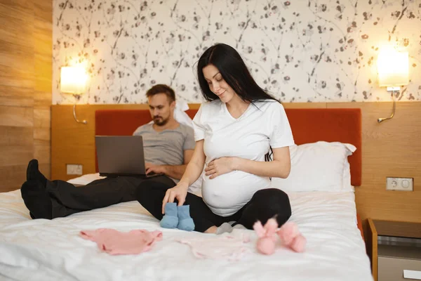 幸せなカップル 自宅で新生児のための夫の服への腹のショーを持つ妊娠中の妻 背景に寝室のインテリア 妊娠期間 期待するお母さんとお父さんはベッドで休んでいる — ストック写真