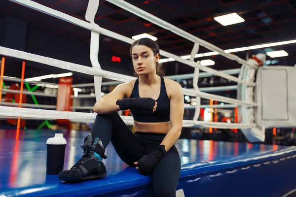 Woman in black boxing bandages sitting at the ropes on the ring, box training. Female boxer in gym, girl kickboxer in sport club, kickboxing workout