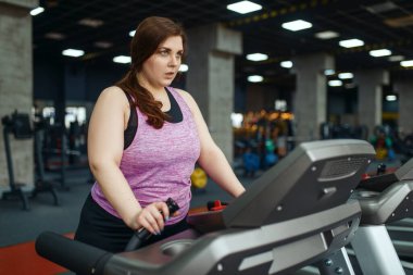 Kilolu bir kadın spor salonunda koşu bandında egzersiz yapıyor, aktif antrenman yapıyor. Obez kadın aşırı kilolarla, obeziteye karşı aerobik egzersizlerle, spor kulüpleriyle mücadele eder.