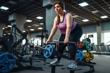 Kilolu bir kadın spor salonunda barla egzersiz yapıyor, aktif antrenman yapıyor. Obez kadın aşırı kilolarla, obeziteye karşı aerobik egzersizlerle, spor kulüpleriyle mücadele eder.