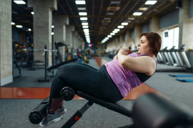 Kilolu bir kadın spor salonunda dambıllarıyla basın üzerinde egzersiz yapıyor, aktif antrenman yapıyor. Obez kadın aşırı kilolarla, obeziteye karşı aerobik egzersizlerle, spor kulüpleriyle mücadele eder.