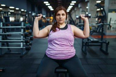 Kilolu kadın spor salonunda dambıllarla poz veriyor, aktif antrenman yapıyor. Obez kadın aşırı kilolarla, obeziteye karşı aerobik egzersizlerle, spor kulüpleriyle mücadele eder.