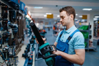 Üniformalı erkek işçi alet deposunda elektrikli testere tutuyor. Hırdavatçıda profesyonel ekipman seçimi, enstrüman süpermarketi