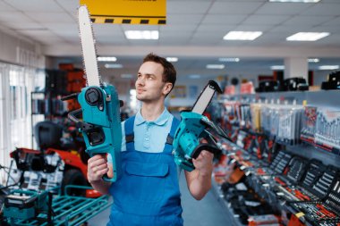 Üniformalı erkek işçi alet deposunda büyük ve küçük testereler tutuyor. Hırdavatçıda profesyonel ekipman seçimi, enstrüman süpermarketi