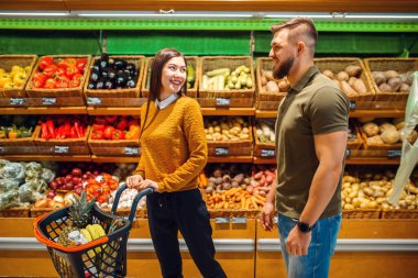Birlikte markette sepeti olan mutlu bir çift. Erkek ve kadın marketten meyve ve sebze alıyor, müşteriler alışveriş yapıyor.