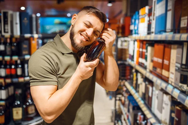 Cheerful man hugs bottle of alcohol in grocery store. Male person buying beverages in market, customer shopping food and drinks