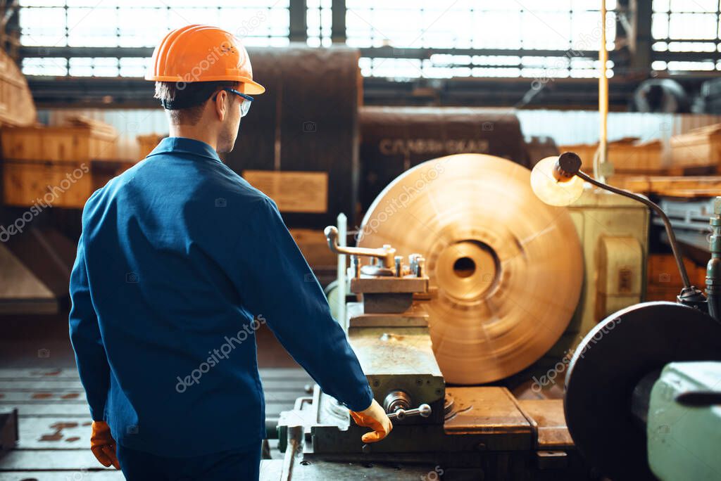 Turner in uniform and helmet works on lathe, factory. Industrial production, metalwork engineering, power machines manufacturing