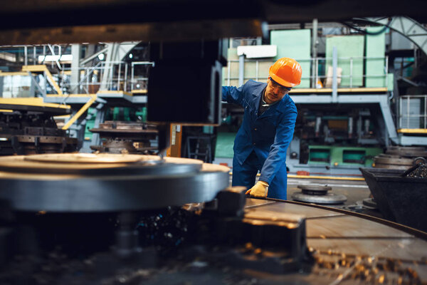 Turner in uniform and helmet stanging at the automated lathe, factory. Industrial production, metalwork engineering, power machines manufacturing