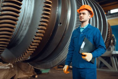 Not defteri olan bir erkek işçi, arka planda büyük bir türbin, bitki. Endüstriyel üretim, metal mühendisliği, güç makineleri üretimi