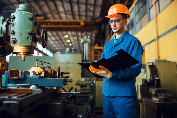 Male worker with notebook, lathe on background, plant. Industrial production, metalwork engineering, power machines manufacturing