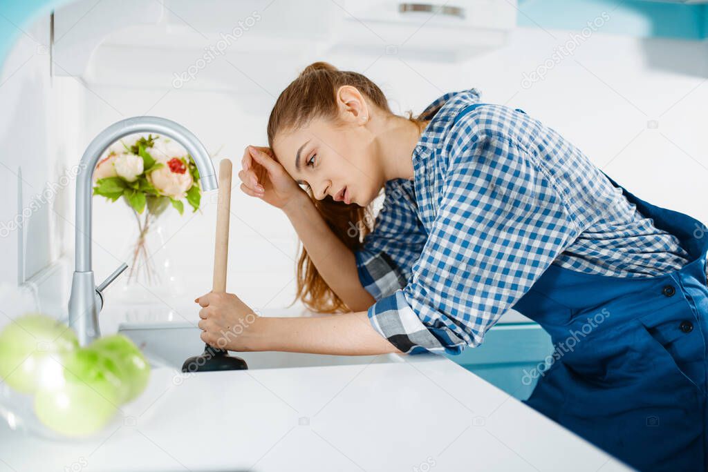 Tired female plumber in uniform holds plunger, clog in the kitchen. Handywoman repair sink, sanitary equipment service at home