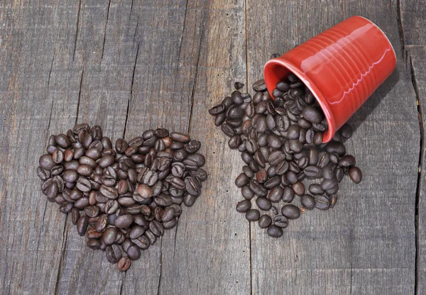 Corazón en forma de granos de café y una taza roja derramada llena de granos en una tabla rústica — Foto de Stock
