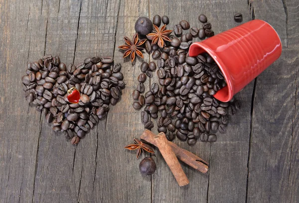 Joya en forma de corazón en granos de café formando un corazón junto a una taza roja derramada llena de granos sobre fondo de madera — Foto de Stock