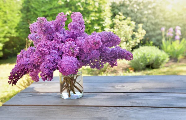 花园里一张木制桌子上的玻璃瓶里放着一束美丽的紫丁香花 — 图库照片
