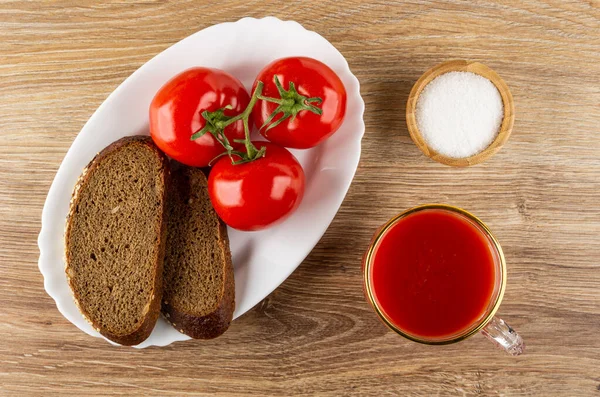Slices of bread, fresh red tomatoes on branch in white dish, salt cellar, cup of tomato juice on wooden table. Top view