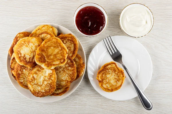 Fried cottage cheese pancakes in glass plate, bowls with strawberry jam, sour cream, fork and pancake in white plate on wooden table. Top view