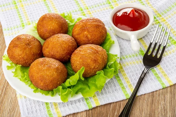 Small round fried pies on leaves lettuce in white glass plate, sauceboat with ketchup, fork on checkered napkin on wooden table