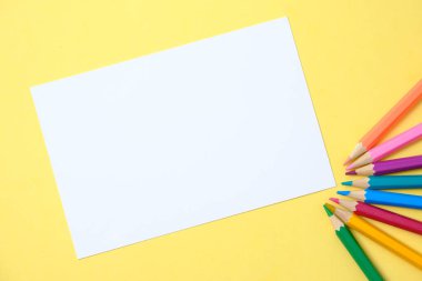 Sarı arka planda beyaz kağıt ve renkli kalemler.