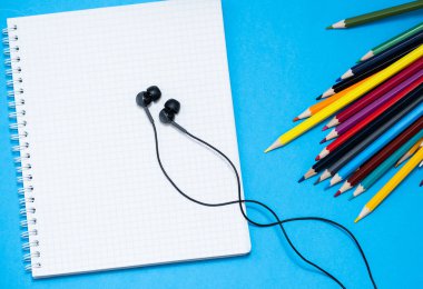 Çizim için renkli kalemler, kulaklıklar ve mavi arka planda bir defter.