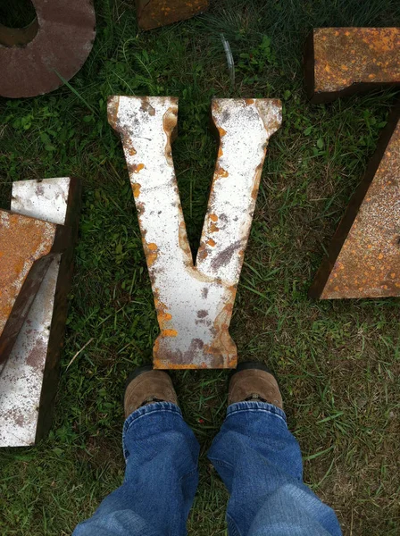 Legs standing over a rusty metal letter "V" — Stock Photo, Image
