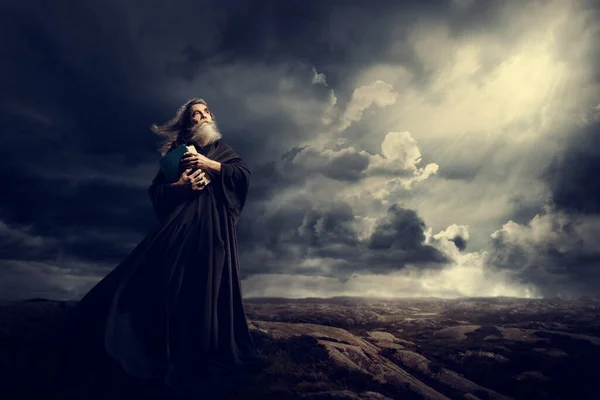 モンク ホールディング聖書 嵐の山の黒いローブの古い司祭 神の空の光を見上げて — ストック写真