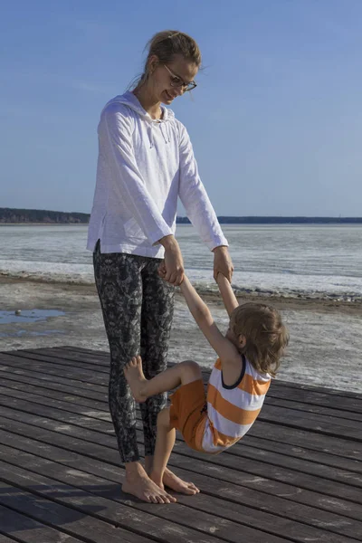 Junge Mutter mit ihrem Kind, das Händchen hält und am Strand Sport treibt. — Stockfoto