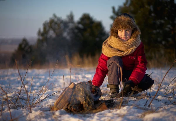 Çocuk tarafından yangın ve İçecek sıcak çay kışın karlı ormanda oturur. Gün batımında.