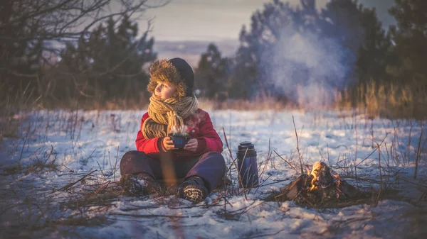 Çocuk tarafından yangın ve İçecek sıcak çay kışın karlı ormanda oturur. Gün batımında.