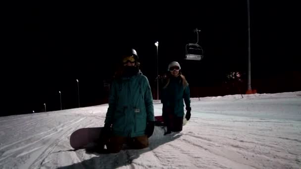 's Nachts paardrijden op het skigebied. Twee vrouwelijke snowboarder staan op de knieën aan de voorkant van de camera en klaar om te rijden. Meisjes gekleed in snowboard apparatuur wachten op start rit — Stockvideo