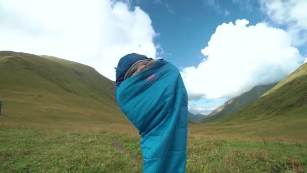 Young woman jumping in sleeping bag at sunset in the forest. Girl dances in sleeping bag on the magnifiscant green meadow between mountain. — ストック動画