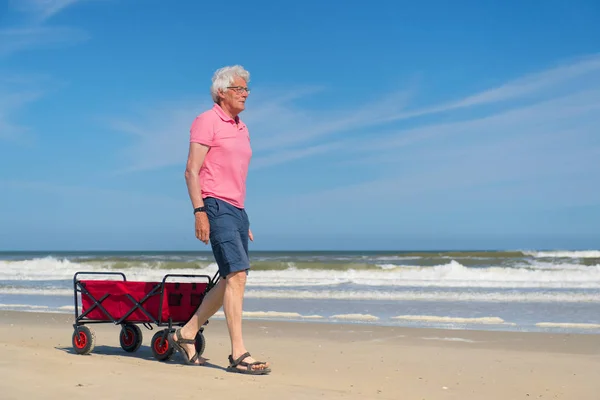 Senior mannen gå med röda vagn på stranden — Stockfoto