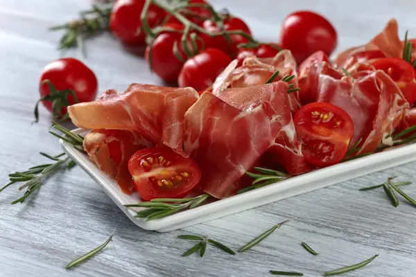 Prosciutto com alecrim e tomate em uma mesa de madeira — Fotografia de Stock